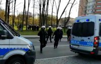 Umundurowani policjanci przebiegają przez ulicę, widoczne dwa zaparkowane radiowozy.