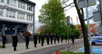 Policjanci wraz z funkcjonariuszami Straży Pożarnej oraz Straży Miejskiej oddają honor przed budynkiem Komendy Miejskiej Policji w Tychach, widoczny policyjny radiowóz.