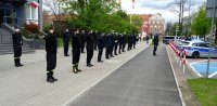 Policjanci wraz z funkcjonariuszami Straży Pożarnej oraz Straży Miejskiej oddają honor przed budynkiem Komendy Miejskiej Policji w Tychach, widoczny policyjny radiowóz oraz wóz funkcjonariuszy Straży Miejskiej.