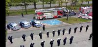 Policjanci wraz z funkcjonariuszami Straży Pożarnej oraz Straży Miejskiej oddają honor przed budynkiem Komendy Miejskiej Policji w Tychach, widoczny policyjny radiowóz oraz wozy funkcjonariuszy Straży Pożarnej.