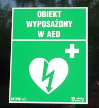 Zielona tabliczka z oznaczeniem &quot;Obiekt wyposażony w AED&quot; serce z błyskawicą oraz krzyżyk.