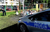 Dwóch umundurowanych policjantów stoi przed grupą dzieci i rozmawia z nimi. Dzieci siedzą na gumowym podłożu, świeci słońce. Widoczny policyjny radiowóz.