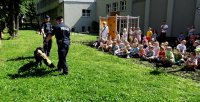 Dwóch umundurowanych policjantów stoi przed grupą dzieci, pomiędzy nimi znajduje się pies służbowy.