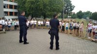 Dwóch umundurowanych policjantów prowadzi prelekcję z dziećmi, które stoją naprzeciwko.