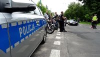 Policyjny radiowóz, zaparkowane motocykle, stojący obok motocykliści, z prawej strony widoczny policjant, a na drodze zatrzymany czarny samochód.