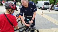 Policjant pomaga chłopakowi zapiąć lampkę na rower.