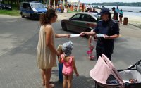 Umundurowana policjantka rozmawia z kobietą, która stoi z dziećmi i daje jej książeczkę z zasadami bezpiecznej kąpieli.