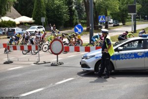 Policjant wydziału ruchu drogowego stoi obok radiowozu i zabezpiecza wyścig kolarski.