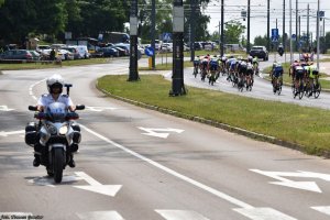 Na jezdni widoczny policjant na motocyklu, po przeciwnej stronie jezdni kolarze uczestniczący w wyścigu.