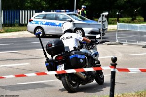 Policjant wydziału ruchu drogowego na motocyklu, widoczny policyjny radiowóz.