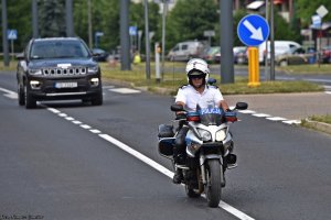 Policjant wydziału ruchu drogowego na motocyklu jedzie przed czarnym samochodem.