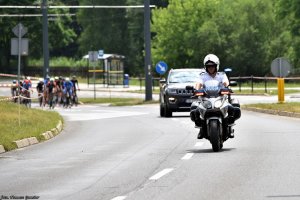 Policjant na motocyklu za nim samochód, a dalej rowerzyści.