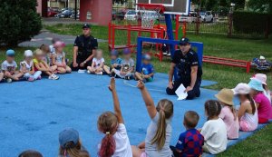 Dzieci siedzą dookoła niewielkiego boiska, dwaj umundurowani policjanci siedzi wśród nich i rozmawiają o bezpieczeństwie, dzieci głoszą się do odpowiedzi.