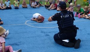 Dzieci siedzą dookoła niewielkiego boiska, dwaj policjant tłumaczy jak zachować się podczas ataku zwierzęcia, na środku skulona dziewczynka.