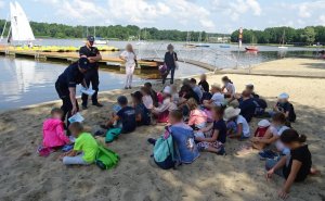 Grupa dzieci siedzi na plaży, przed nimi policjantka rozdaje edukacyjne książeczki obok stroi policjant i dwie opiekunki.