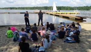 Grupa dzieci siedzi na plaży, przed nimi stoi policjant i policjantka i rozmawiają o bezpieczeństwie dzieci nad wodą. W tle widoczne jezioro oraz molo.