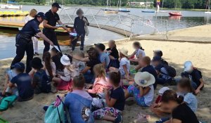 Grupa dzieci siedzi na plaży, przed nimi policjant i policjantka rozdają edukacyjne książeczki.