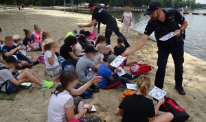 Policjanci rozdają edukacyjne książeczki dzieciom, które siedzą na piasku na plaży.