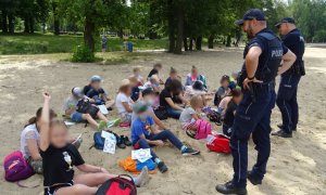 Grupa dzieci siedzi na plaży, przed nimi stoją policjanci i rozmawiają o bezpieczeństwie.