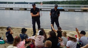 Policjanci stoją przed grupą dzieci, które siedzą na piasku i rozmawiają na temat bezpiecznej kąpieli, za nimi widoczne jezioro.