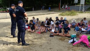 Policjanci stoją przed grupą dzieci, które siedzą na piasku.