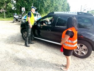 Policjant rozmawia z kierowca samochodu obok stoi pani pracownik Polskich Linii Kolejowych, w tle widoczny radiowóz.