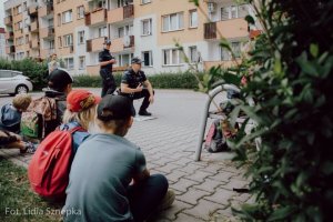 Umundurowany policjant i policjantka rozmawiają z grupą dzieci, które siedzą na trawie.