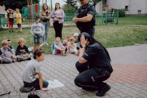 Policjantka kuca przed chłopcem i zasłania rękami głowę, obok widoczny policjant i inne dzieci.