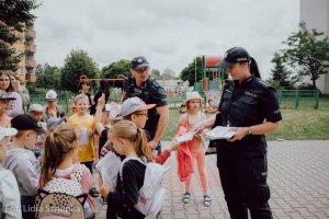 Policjantka rozdaje dzieciom kolorowanki obok drugi policjant.