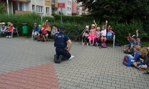 Policjant rozmawia z dziećmi, które siedzą przed nim na ławkach.