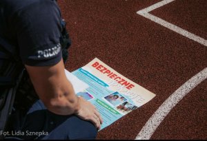 ramię policjanta w mundurze, na ramieniu widoczny jest napis POLICJA przed sobą ma plakat dotyczący bezpiecznych wakacji