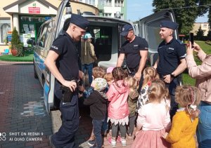 Umundurowani policjanci stoją z tyłu radiowozu, do którego wchodzą dzieci.