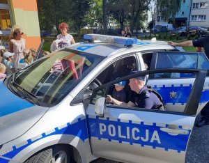 Dzieci wewnątrz radiowozu obok policjant, widoczne inne osoby.