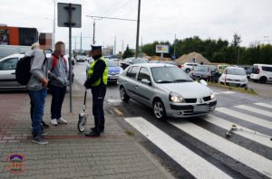 Policjant rozmawia z osobami w ramach przeprowadzanej symulacji wypadku, na pasach leży hulajnoga przed nią samochód.