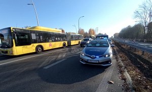 Widoczny zółty autobus komunikacji miejskiej oraz policyjny radiowóz, pomiędzy jedzie samochód.