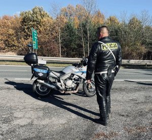 Policjant Wydziału Ruchu Drogowego przy drodze przed nim policyjny motocykl.