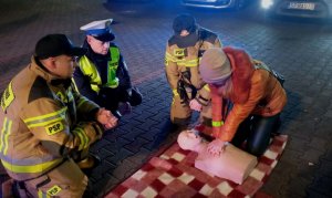 Dwóch strażaków i policjant obok kobieta ćwiczy pierwszą pomoc na manekinie.