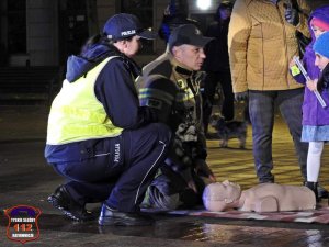 Policjantka obok strażak, który tłumaczy pierwszą pomoc kobiecie z dzieckiem.