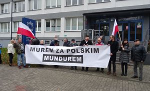 Grupa osób stoi przed budynkiem komendy i trzyma baner z napisem &quot;Murem za Polskim Mundurem&quot;.