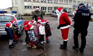 Dzieci z czapeczkami mikołaja, mikołaj, obok umundurowany policjant i mężczyzna z czerwoną czapką. Widoczny policyjny radiowóz.