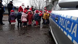 Widoczny napis &quot;Policja&quot; z radiowozu, dalej dzieci z czerwonymi czapeczkami, mikołaj, policjanci i strażnicy miejscy.