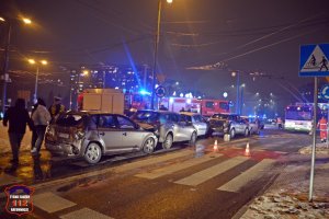 Na zdjęciu widoczny wóz straży pożarnej, policjanta, pojazdy które uczestniczyły w stłuczce, osoby postronne, jest ciemno.