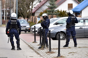 Trzech policjantów w umundurowaniu zabezpieczającym mecz piłki nożnej, w tle samochody osobowe.