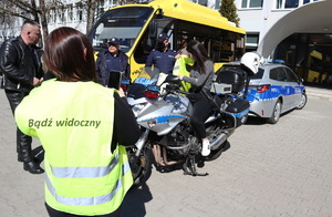 Dziewczyna z odblaskową kamizelką z napisem na plecach &quot;Bezpieczny pieszy&quot; robi zdjęcie dziewczynie na policyjnym motocyklu. widoczni umundurowani policjanci.