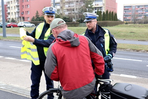 Dwóch umundurowanych policjantów z wydziału ruchu drogowego rozmawia z rowerzystą, przekazują mu kamizelkę odblaskową.