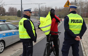 Rowerzysta zakłada kamizelkę odblaskową, obok niego dwaj umundurowani policjanci, w tle radiowóz.