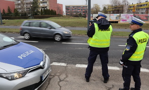 Umundurowani policjanci wydziału ruchu drogowego stoją przez radiowozem, jeden z nich mierzy dopuszczalna prędkość pojazdów.