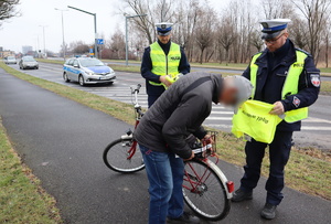 Rowerzysta zakłada na rower opaskę odblaskową obok stoi dwóch umundurowanych policjantów, za nimi policyjny radiowóz.