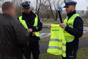 Policjant drogówki trzyma kamizelkę odblaskową z napisem &quot;Bądź widoczny&quot; obok stoi drugi policjant oraz mężczyzna, który otrzymał kamizelkę.