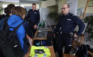 Dwóch umundurowanych policjantów stoi przy stoliku z gadżetami i ulotkami i rozmawiają z młodzieżą.